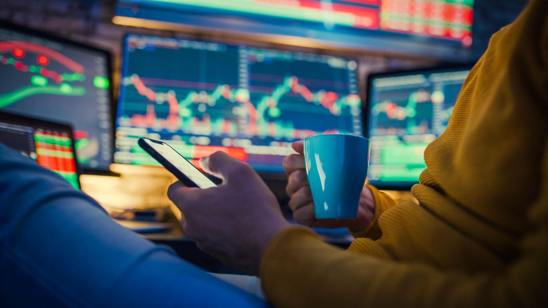 Man using mobile phone and having coffee while monitoring stock market on computer screens at desk